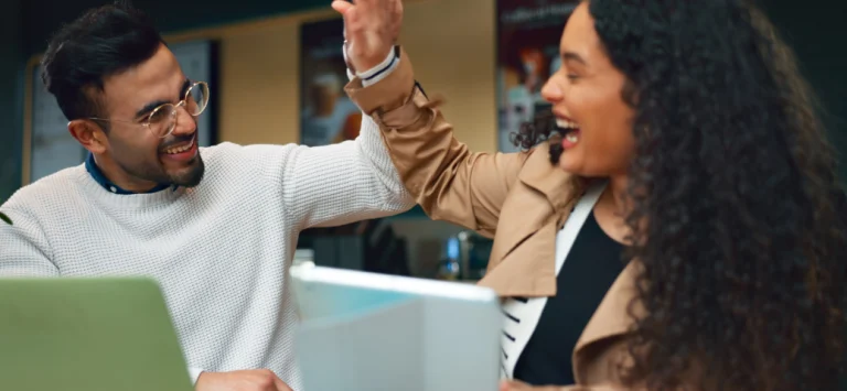 Colegas de trabalho sorriem ao celebrar com um "high five" em mesa com notebook e tablet em um ambiente corporativo.